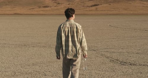 An Adult Young Man Walks Barefoot Along the Bottom of a Dry Lake Among Hills with Dried Grass and