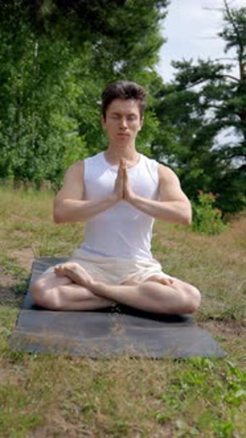 Vertical of Young Athletic Man Meditates in Lotus Pose on Grassy Meadow