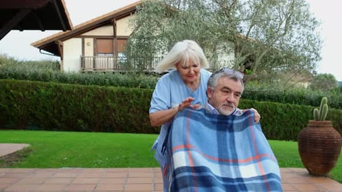 Nurse covering elderly man in wheelchair with blanket in garden