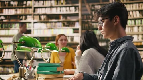 Portrait of Young Man with Glasses Reading Open Book at Wooden Library Table
