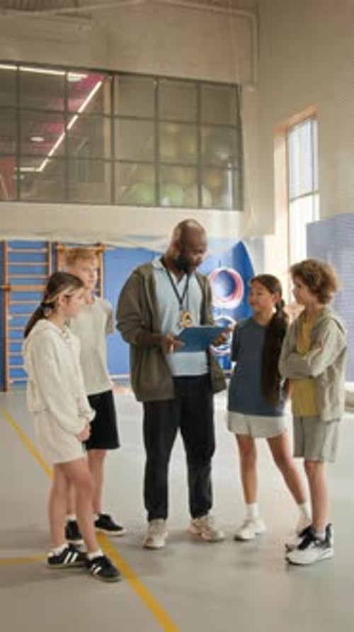 Black Man as Coach Talking to Children in Gym and Holding Clipboard
