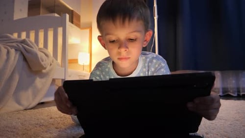 Porträt eines Jungen, der im Schlafzimmer auf einem Teppich liegt und nachts Spiele auf einem Tablet-Computer spielt