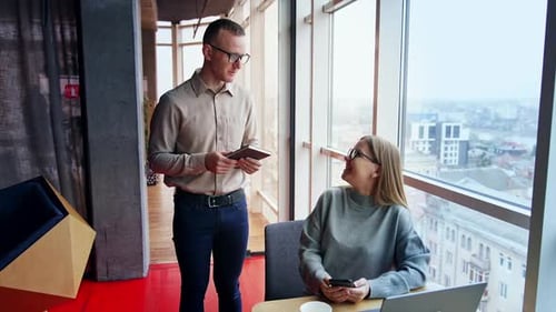 Relaxed smiling people in the office. Lady sits at desk looking up at man standing beside.