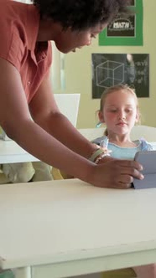 Vertical video: Approaching teacher in rust shirt guiding child's hands to use tablet at classroom