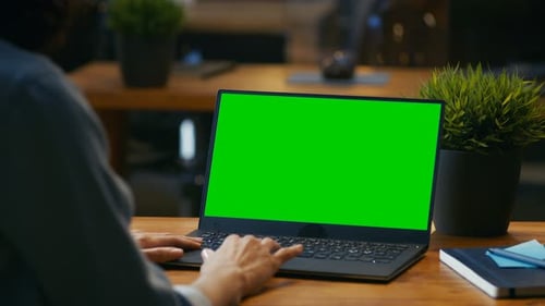Female Office Worker at Her Desk Works on a Laptop with Mock-up Green Screen. Over the Shoulder Foo