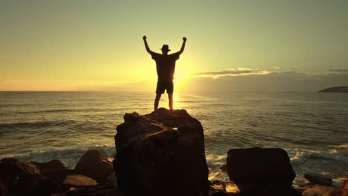 The Silhouette of a Man in a Hat Stands Atop a Large Oceanfront Rock Against a Breathtaking Sunrise