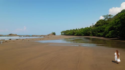 Woman walking on wide empty wet sand beach with rocks and palms on clear morning