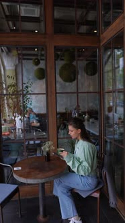 A Vibrant Young Woman Enjoys Coffee on the Café's Summer Terrace Holding a Smartphone in Her Hand