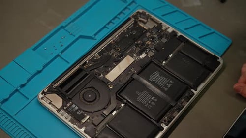 Techniker untersucht den zerlegten Laptop mit Handschuhen, überprüft die interne Motherboard-Batterie und