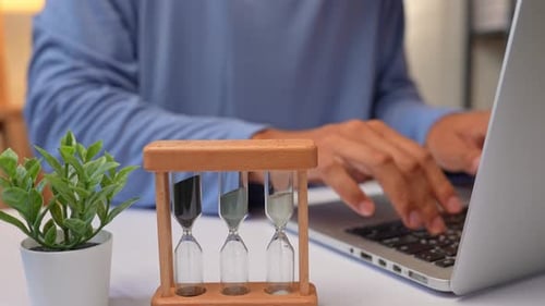 A person works at a laptop with an hourglass beside them.