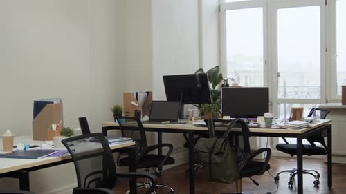 Messy Office Workspace with Computers, Papers, Tables and Chairs near Window