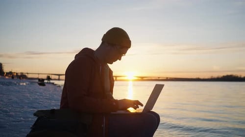 Young Adult Using Laptop by River at Sunrise