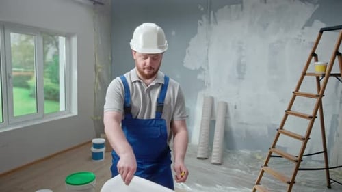 Männlicher Vorarbeiter schaut sich ein großes Blatt mit einem Wohnungsplan an und denkt über ein Reparaturprojekt nach