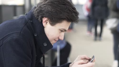 Young, Teenage Boy Playing Game on Smartphone by Tram Stop in City Alone