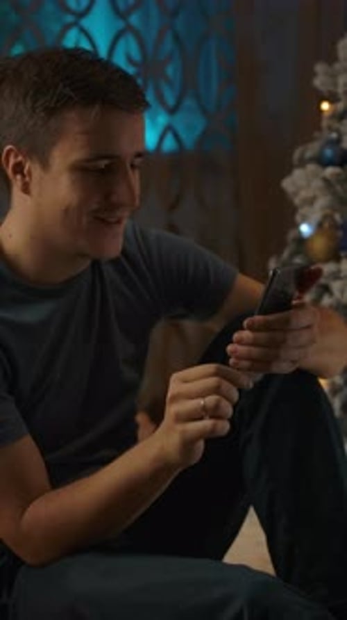 Vertical of Young Caucasian Man Using Phone Sitting Near Cozy Christmas Tree December