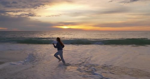 Silhouette of a Woman Running Along the Ocean Coast Barefoot on a Breaking Wave at Sunset The