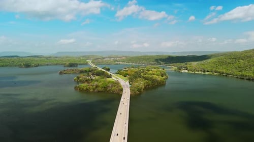 Lange Autobahnbrücke, die sich über einen ruhigen Fluss und eine Insel erstreckt, umgeben von üppigen grünen Hügeln, weich