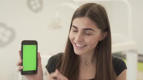 Close-up view of a young woman holding green screen smartphone, showing her thumb up and smiling.
