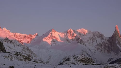 Snow-covered mountain peaks glow pink at sunrise under a clear sky, panning scenic landscape shot