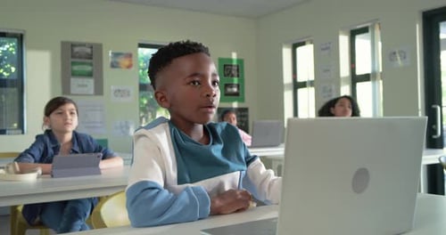 African American boy at desk raising hand, responding to teacher, using silver laptop