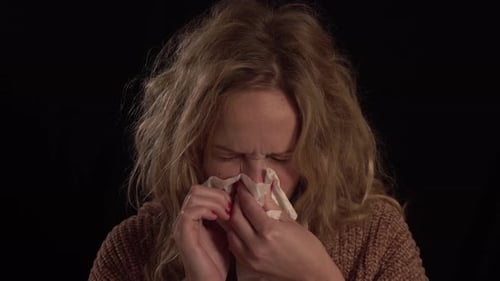 Une jeune femme blonde européenne se mouche. Elle a le nez qui coule et elle se sent très malade.
