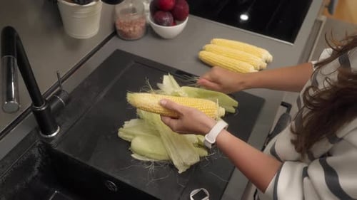 Woman deshusking ear of corn in bright kitchen