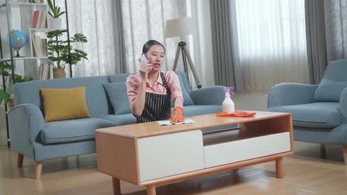 Asian Female Housekeeper With An Apron Talking On Smartphone While Cleaning The Table By The Spray