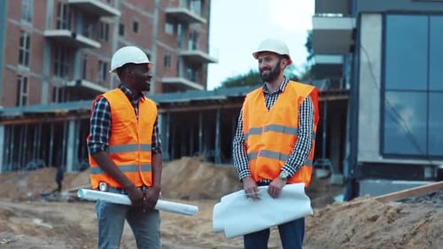 Two Multiracial Construction Workers in Safety Helmets Holding the Building Blueprints and Actively