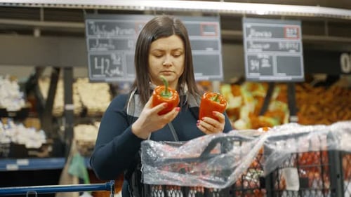 Girl with a Trolley in a Supermarket Chooses Fresh Red Pepper