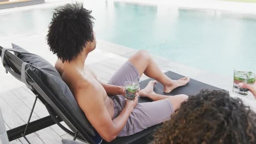 Video of happy diverse couple on holiday relaxing with drinks in sun and talking by swimming pool