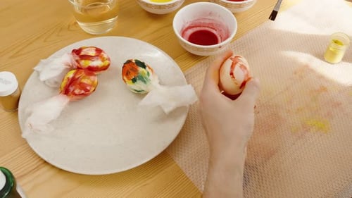 Hand hält ein verziertes Osterei, mit eingewickelten Eiern und Schalen voller Farbe auf einem Tisch, Vorbereitung für
