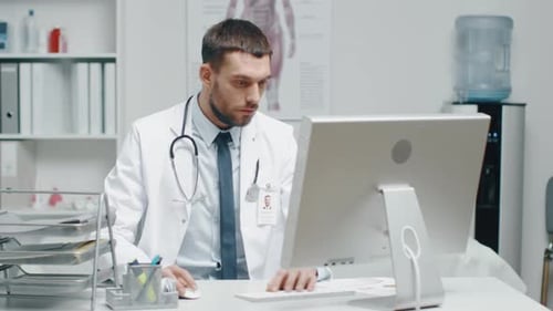 Close-up of a Male Doctor is Working at His Desk. He Uses Personal Computer and Consults Documents.