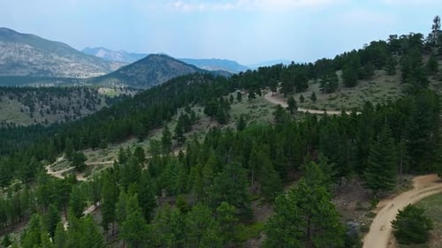 Aerial view of a winding mountain road leading through green forests and rocky terrain, with distant