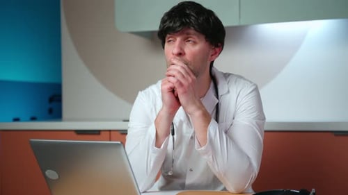 A Thoughtful Serious Male Doctor is Sitting at a Table Using a Laptop Immersed in His Thoughts