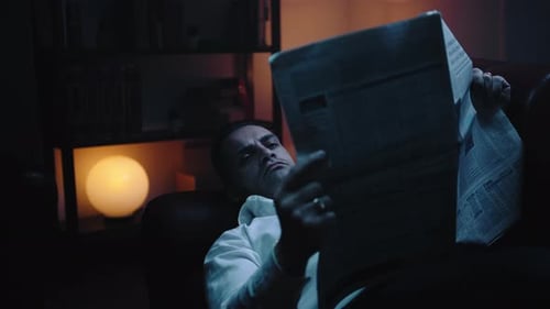 Businessman Lying on Sofa Reads the Newspaper at Night