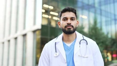 Portrait of a serious doctor in white coat looking at camera, standing outside near hospital