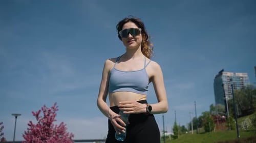 Athletic Woman Drinking Water Recovering After Training Session in Sunlit City Park Sportswear