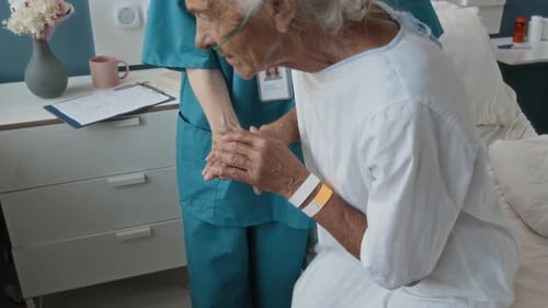 Elderly Woman Standing up from Bed with Assistance of Young Doctor in Hospital
