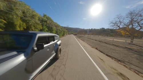 Vista FPV inversa de un todoterreno que corre por una carretera rural aislada durante un día soleado.