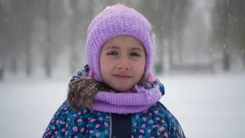 A Little Girl Stands Under Snowfall in the Park A Girl in a Jacket and Hat Looks at the Camera