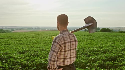 Back View of a Farmer Wearing a Plaid Shirt Standing on the Field A Man Holding a Spade Looking at
