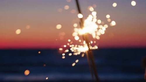 Sparklers Held By Hands Against a Colorful Sunset Over the Ocean