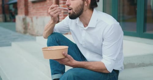 Businessman lunch eating salad in the city street near office building. Cheerful bearded man having