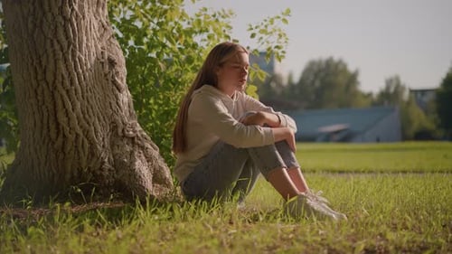Thoughtful Woman Sitting on Grass in Sunlight with Urban Stadium in Background