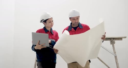 Asian woman engineer checks laptop while male contractor reads large blueprint, coordinating