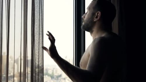 Der Mann im Handtuch enthüllt den Vorhang und bewundert die Aussicht vom Fenster zu Hause
