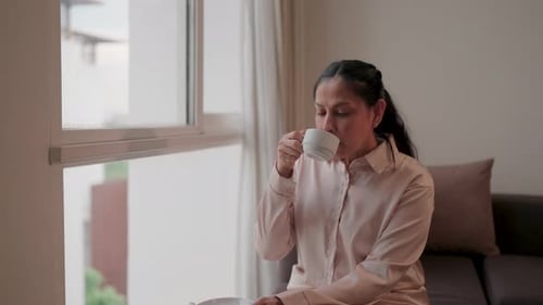 Middle-aged woman is sipping a warm drink by the window where sunlight streams