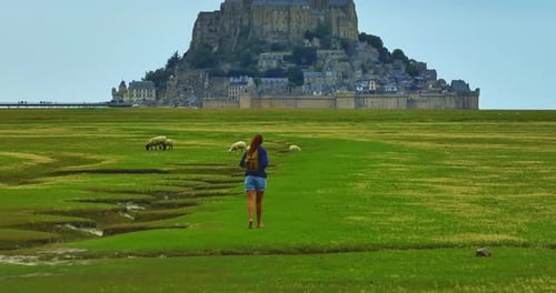 A Female with a Backpack Visits the Mont Saint Michel Castle While Walking Through Green Meadows