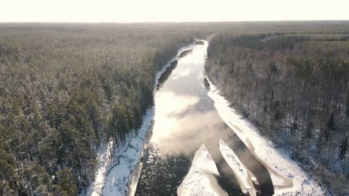 Aerial view of a winding river through a dense snow-covered forest in winter, with a curving road ru