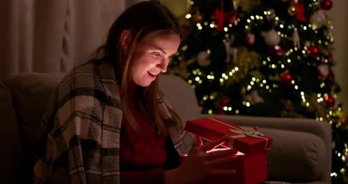 Woman Opens Christmas Present near Sparkling Tree